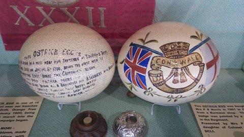 Two colourful and highly decorated hand-painted Ostrich eggs sit in a display case surrounded by smaller artefacts. The eggs on the right has the DCLI insignia and the egg on the left is decorated in handwritten script